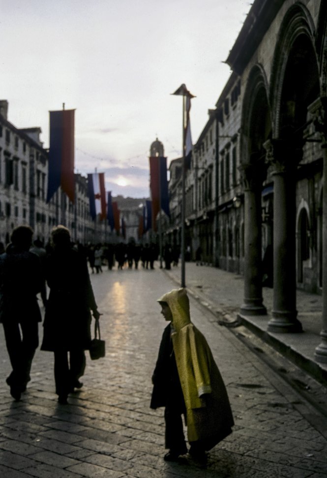 Free image of Child standing in a rain coat in front the setting sun on a rainy brick street, Europe