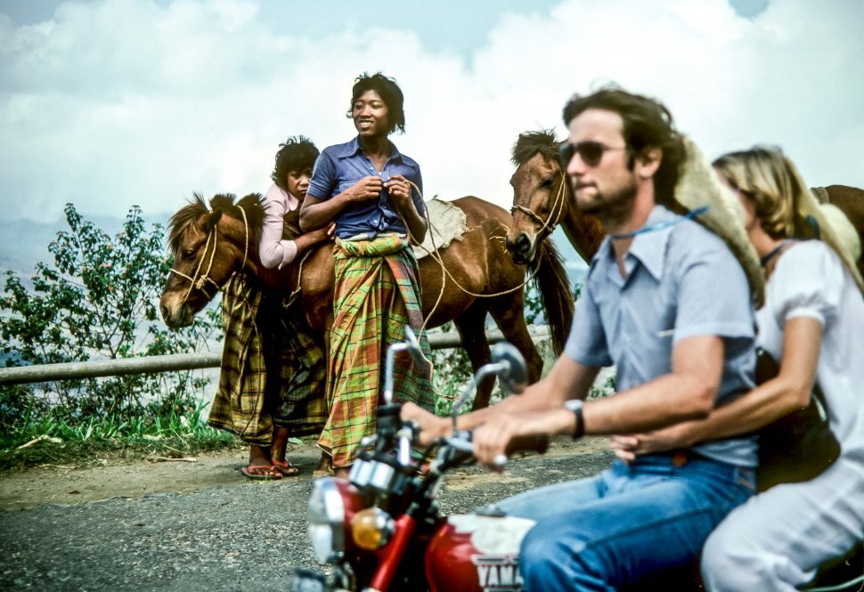 Free image of Couple riding a motorcycle past locals walking with their horses.