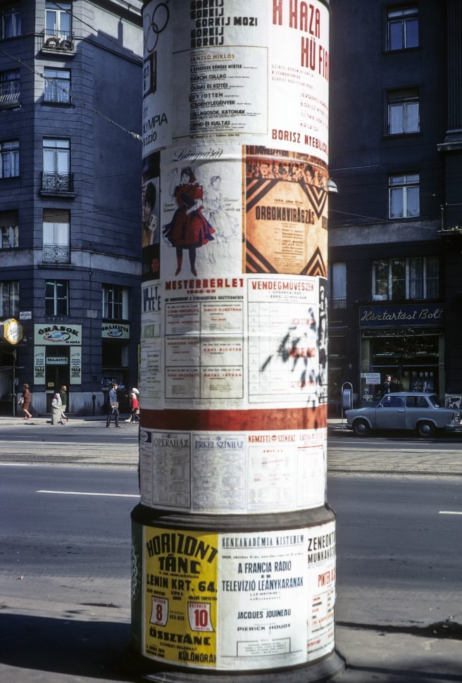 Free image of Posters on a city light post with pedestrians walking behind it.